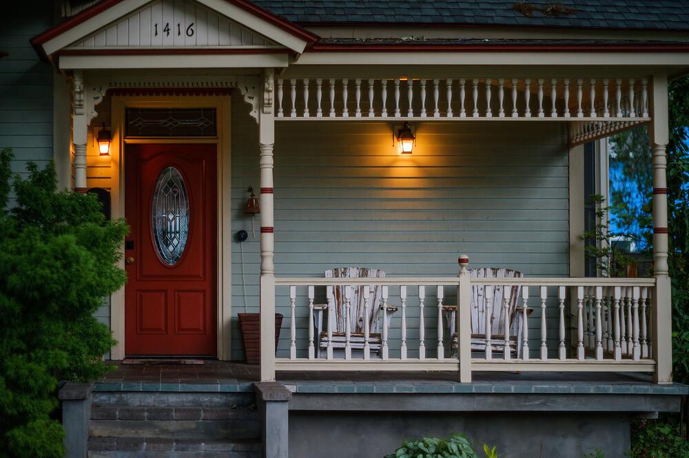 A covered front porch with wooden chairs, a red door and warm evening lighting