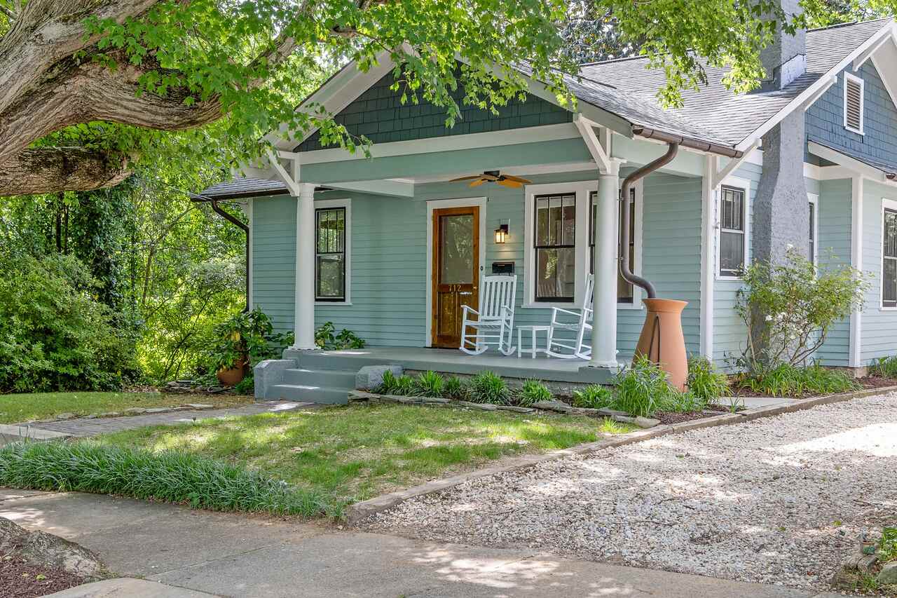 A small bungalow with a low porch and rocking chairs surrounded by heavy planting