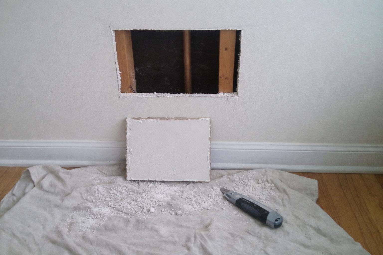 A small rectangular access hole cut into an interior drywall wall with the removed square leaning below it