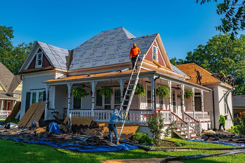 Two workers on ladders at the roofline of an older wood-frame house with a deep porch
