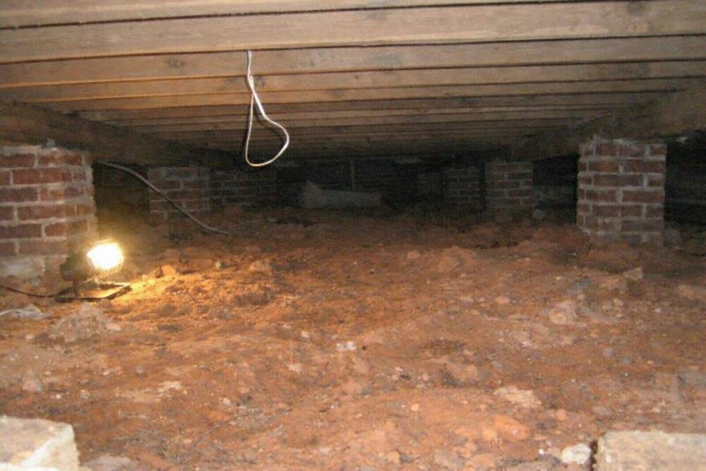Bare red clay soil under low floor joists inside a crawl space with no vapor barrier